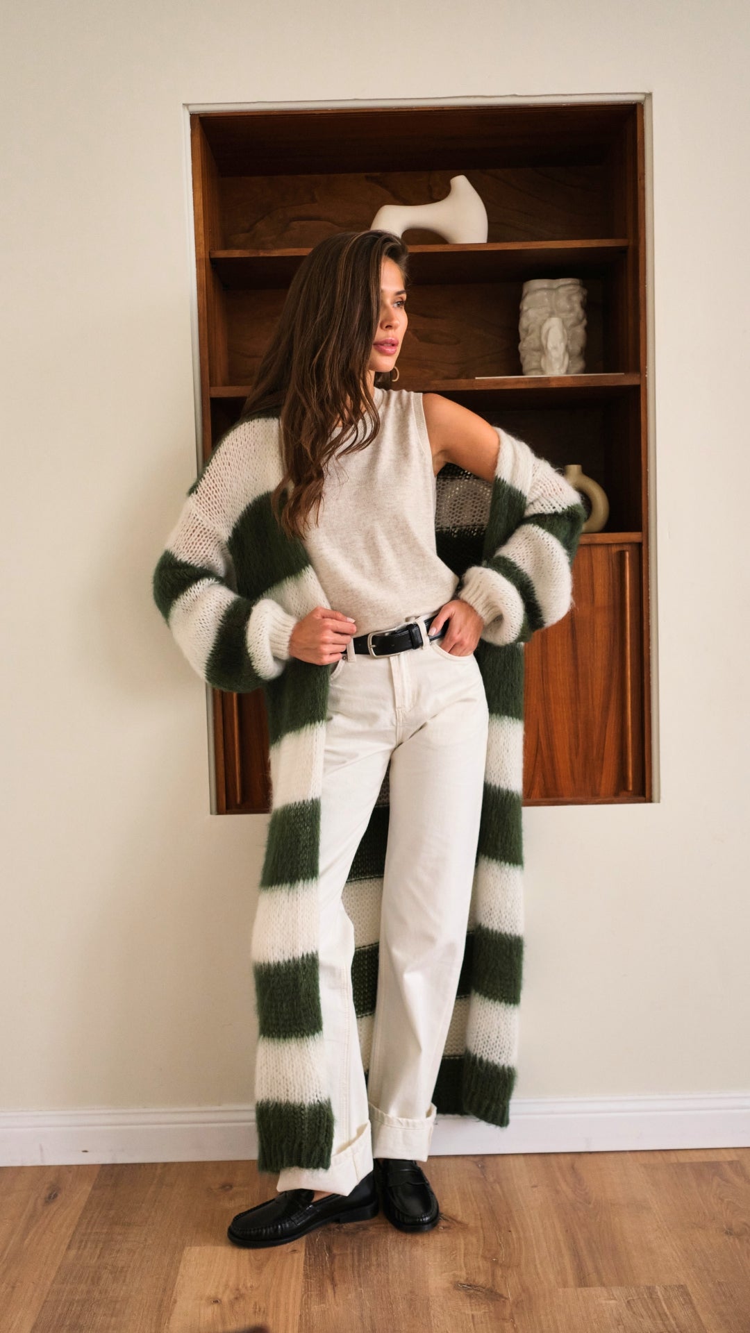 sleeveless beige top tucked into white high-waisted trousers with a black belt, styled with a long green and white striped knit cardigan and black loafers